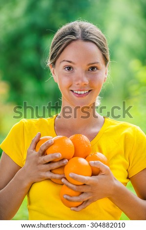 Portrait of dark-haired smiling beautiful young woman in yellow blouse with oranges, against green of summer park.