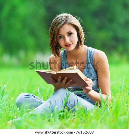 against background of green meadow (grass meadow) sat young, charming girl - student reads book