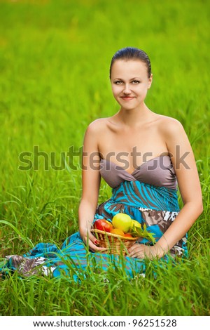 Portrait of beautiful young smiling woman holding a basket of fruit sits on grass at summer green park