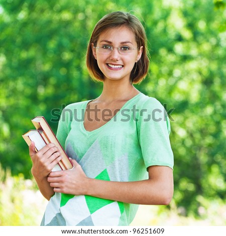 Portrait of pretty, slender student in glasses holding book against background of summer green park