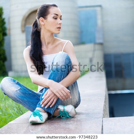 Young beautiful dark-haired woman sits on stone fencing about many-storeyed building.