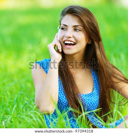 Young beautiful woman in blue dress sitting on grass in summer city park and talking on cell phone.