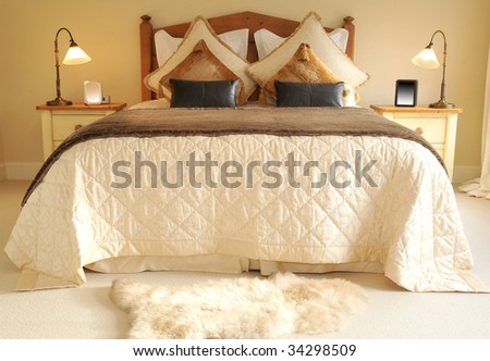 Viewed from the end a bed in a bedroom with bedside cabinets, lights and large billows and cushions
