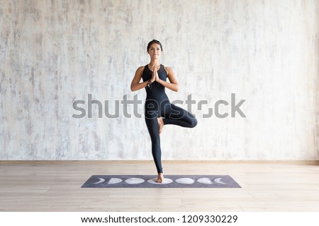 Beautiful young brunette woman yoga instructor doing vrikshasana on a mat in a wooden floor standing in the gym with day lighting