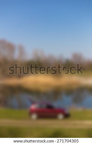 Blurred background red car with an open trunk along the narrow river near a dirt road. The opposite bank with reeds, deciduous trees without leaves. Spring, blue sky, young grass, sunny day.