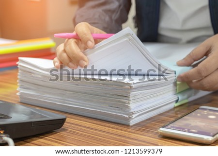 Teacher hand is searching for checking student homework assignments on desk in school office. Unfinished paperwork stacked in archive with color papers. Education and business concept.