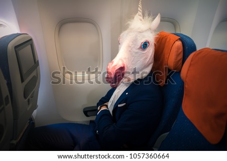 Young funny man in comical mask travels by plane. Unusual passenger in elegant suit sits alone inside the aircraft and ready to take off. Freaky traveler in air voyage. Unicorn enjoys his flying