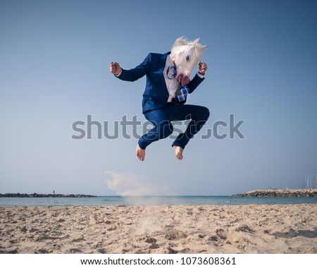 Stylish man in funny mask and suit jumps on beach. Unicorn have fun on the shore and enjoys vacation. Elegant traveler on background of sea and sky