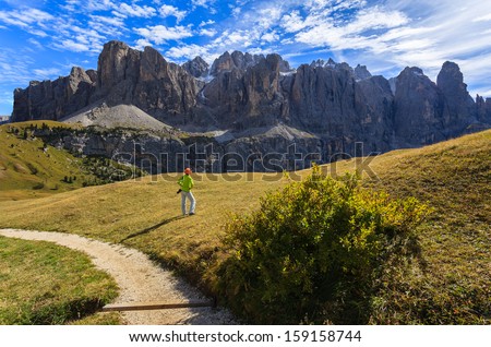 Young woman photographer standing with camera hanging off shoulder at mountain alpine valley trail white clouds blue sky near Passo Gardena, Dolomites Mountains, Italy