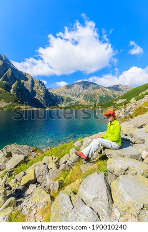 Young woman tourist sits on a rock and looks at mountain lake Czarny Staw on trail from Morskie Oko, High Tatra Mountains, Poland
