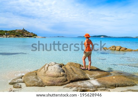Young woman tourist standing on a rock and looking at crystal clear turquoise sea water at Punta Molentis beach, Sardinia island, Italy