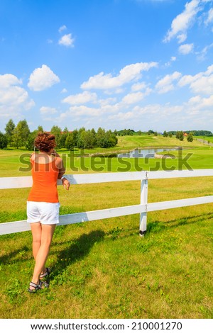 Young woman stands against a white fence and looks at golf club area on sunny summer day, Poland