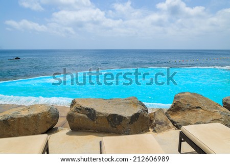 Sunbeds with lava rocks and turquoise water of infinity swimming pool at coast of Atlantic Ocean, Madeira island, Portugal