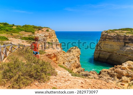 Young woman tourist standing on cliff rock and looking at sea on coast of Portugal, Algarve region