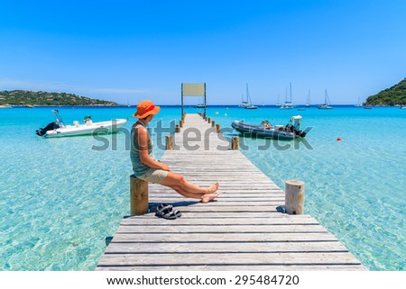 Young woman tourist sitting on wooden jetty in Santa Giulia marina with turquoise sea water, Corsica island, France