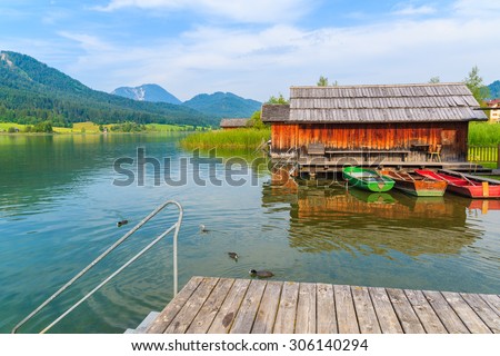 Wooden jetty and fishing boats with wooden houses on shore of Weissensee lake in summer landscape of Carinthia land, Austria