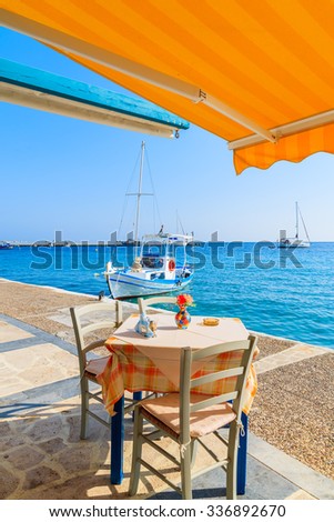 Table with chairs in Greek tavern on coast of Samos island with fishing boat in distance, Kokkari town, Greece