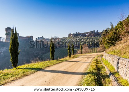Cypress trees bordering a walk path to a medieval fortress and a sanctuary with steeple on misty hills in a contryside of bushes and cypress trees in a winter sunny day