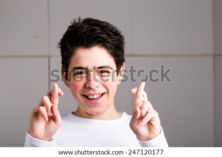 Latin young boy with acne in a white long sleeve t-shirt smiles crossing fingers of both hands as superstitious  gesture to get luck on an industrial background