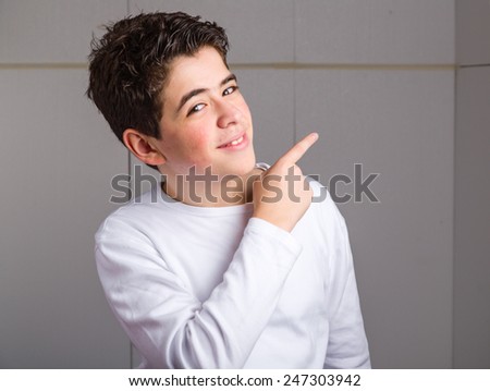 Caucasian young boy with acne in a white long sleeve t-shirt pointing up to his left side with right forefinger  on industrial background