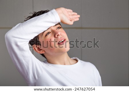 Cute Caucasian boy with acne-prone skin in a white long sleeved t-shirt holding his right arm on the front as protecting from sun or wiping the sweat