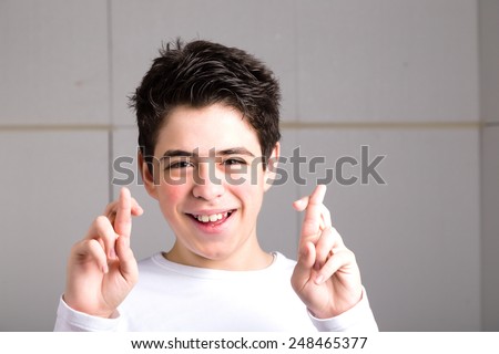 Latin young Smooth-skinned boy in a white long sleeve t-shirt smiles crossing fingers of both hands as superstitious  gesture to get luck on an industrial background