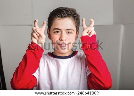 Caucasian smooth-skinned boy in a red and white long sleeve t-shirt raising hands bends index and middle fingers as quotes gesture on industrial background
