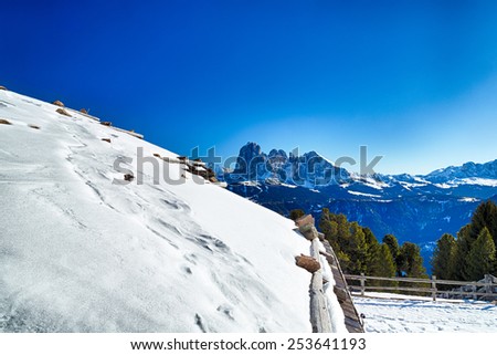 Roof of alpine chalet surrounded by a fence in the snow in front of a panorama of snowy peaks on a bright sunny day in winter on Dolomites Alps
