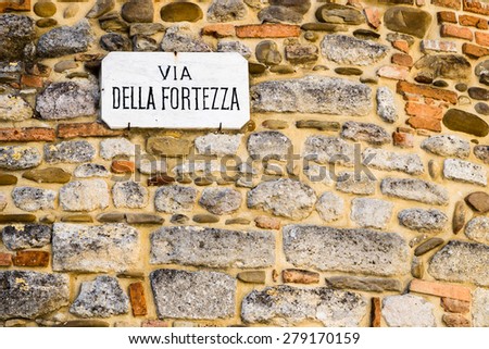 Italian sentence, Via della Fortezza on sign nailed on wall made by smooth stones: meaning is Fortitude road or Fortress road