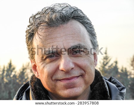 Elegant middle-aged man with green eyes and  salt pepper hair dressed in dark blue padded jacket is smiling reassuringly in the countryside