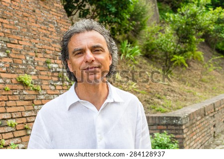 Handsome tanned middle-aged man with salt pepper hair and green eyes dressed with linen white shirt and blue pants in Italian medieval outdoors: he is relaxed near brick walls of a medieval building