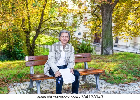 Handsome 50 years old man with salt pepper hair and green eyes dressed with grey gabardine, linen white shirt and blue pants is reading documents in a park in Prague