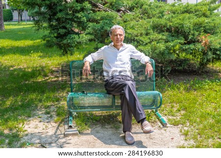 Handsome middle-aged man with salt pepper hair dressed with white shirt, blue slacks and beige moccasins is resting on a bench in city park keeping his arms opened: he shows a reassuring look