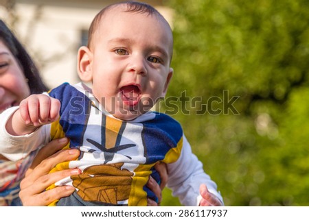 Cute 6 months old baby with Light brown hair in white, blue and brownish long-sleeved shirt is embraced and held by his mother: they seem very happy