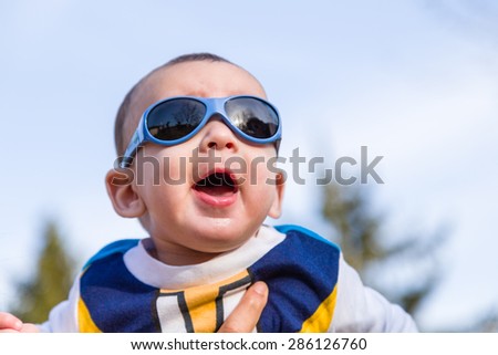 Cute 6 months old baby with Light brown hair in white, blue and brownish long-sleeved shirt wearing blue googles is embraced and held by his mum: he seems very happy and smiles
