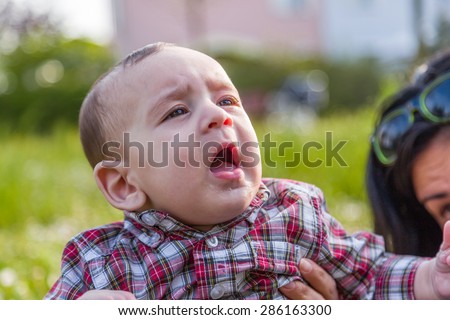 Cute 6 months old baby with Light brown hair in red checkered shirt and beige pants is gaping and looking up while embraced by mother