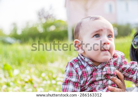 Cute 6 months old baby with Light brown hair in red checkered shirt and beige pants is biting his lips, puffing his cheeks and  looking up while embraced by mother