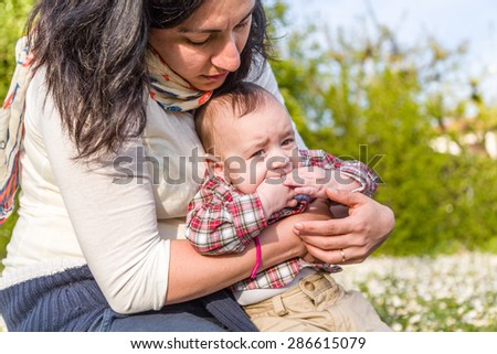 Cute 6 months old baby with Light brown hair in red checkered shirt and beige pants is biting his fingers while embraced by his Hispanic mother