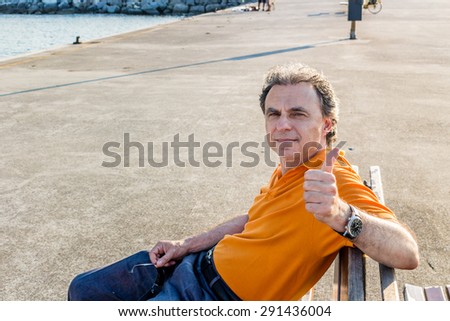 Classy senior sportsman with three-day beard and salt and pepper hair wearing an orange polo shirt while he is sitting on a bench on the pier and showing thumb up