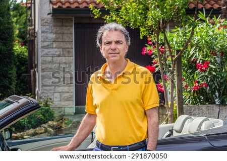 Classy senior sportsman with three-day beard and salt and pepper hair wearing a yellow polo shirt while he is sitting on the door of a dark brown cabriolet car in residential neighborhood