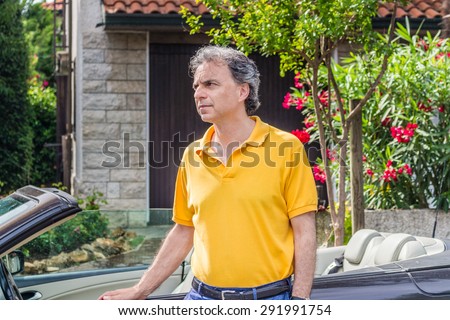 Classy senior sportsman with three-day beard and salt and pepper hair wearing a yellow polo shirt while he is sitting on the door of a dark brown cabriolet car in residential neighborhood