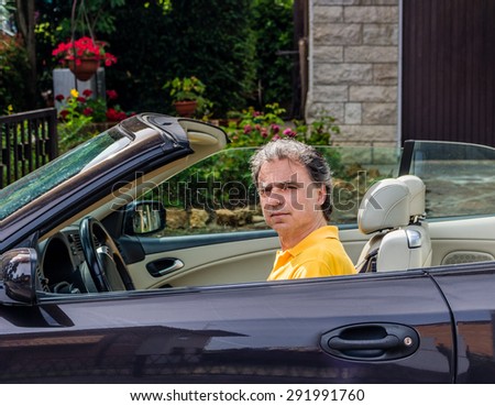 Side view of classy senior sportsman with three-day beard and salt and pepper hair wearing a yellow polo shirt while he is driving a dark brown car in residential neighborhood