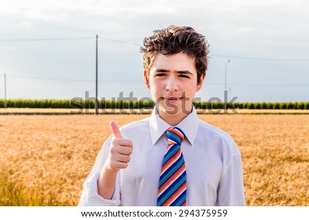 Handsome Caucasian boy wearing a white shirt and a regimental tie with red, fuchsia, orange, blue, indigo and white stripes is making thumbs up sign on a field of golden wheat ears at sunset