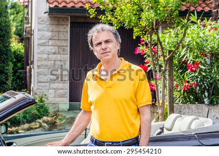 Classy senior sportsman with three-day beard and salt And pepper hair wearing a yellow polo shirt while he is sitting on the door of a dark brown cabriolet car in residential neighborhood