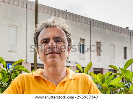 Close up of handsome classy middle-aged sports man with three-day beard and salt cand pepper hair wearing a yellow polo shirt  with old building in the background