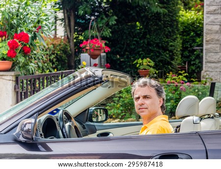 Side view of classy senior sportsman with three-day beard and salt cand pepper hair wearing a yellow polo shirt while he is driving a dark brown car in residential neighborhood