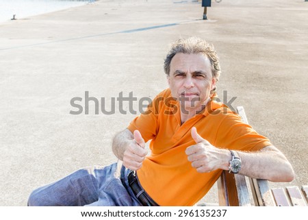 Classy senior sportsman with three-day beard and salt and pepper hair wearing an orange polo shirt while he is sitting on a bench on the pier and showing thumbs up