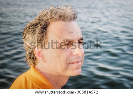 Classy  40 years old sportsman with three-day beard and salt and pepper hair wearing an orange polo shirt while he is thinking in front of the sea