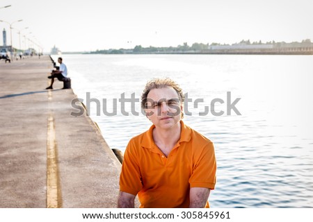 Classy  40 years old sportsman with three-day beard and salt and pepper hair wearing an orange polo shirt while he is sitting on the pier and resting