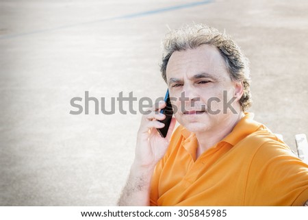 Classy  40 years old sportsman with three-day beard and salt and pepper hair wearing an orange polo shirt while he is sitting on a bench on the pier and talking on a mobile phone